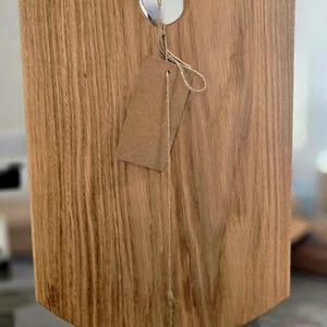 Può includere: Un tagliere in legno color marrone chiaro con una venatura verticale prominente. Il tagliere presenta un intaglio a forma di U nella parte superiore centrale e un bordo inferiore arrotondato. Un cartellino di carta marrone è legato con spago.