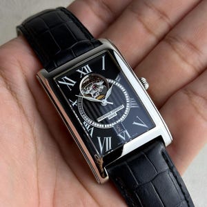 May include: A rectangular silver-tone wristwatch with a black dial featuring Roman numerals, a visible balance wheel aperture, and a black leather strap with a crocodile-embossed texture. The dial displays the text Frederique Constant Geneve.