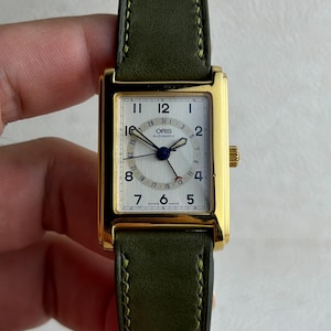 May include: A rectangular gold-tone Oris automatic watch with a white dial, black Arabic numerals, and a date pointer hand. The watch features a textured olive green leather strap with matching green stitching and a gold-tone crown on the side.