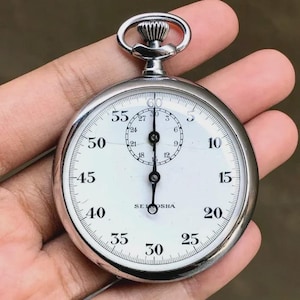 May include: A silver-toned analog stopwatch with a white face, black numerals, and a winding crown at the top. The dial features a main scale from 5 to 60 and a smaller sub-dial marked from 3 to 27. The text SEIKOSHA is printed in black on the face.