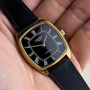 May include: A gold-tone wristwatch with a square-shaped case and a black dial. The watch features Roman numeral hour markers, gold-colored hands, and the text Longines and Automatic on the face. It is attached to a black leather strap.