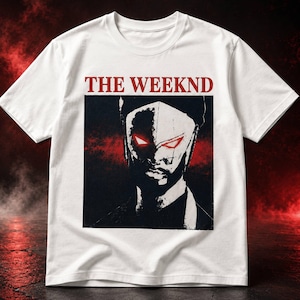 May include: A white short-sleeve t-shirt featuring the text THE WEEKND in red capital letters above a square graphic. The graphic displays a stylized black and white portrait of a person wearing a mask with glowing red eyes against a red background.