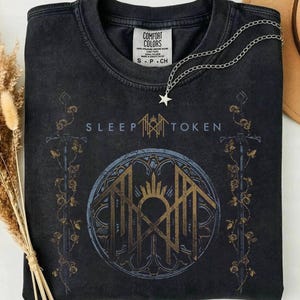Könnte beinhalten: Ein schwarzes T-Shirt mit Rundhalsausschnitt und Comfort Colors Etikett. Das Motiv zeigt den Schriftzug SLEEP TOKEN, ein goldenes Logo in einem kreisförmigen Rahmen sowie zwei vertikale, mit Rosen umrankte Schwerter. Eine silberne Sternkette liegt darauf.