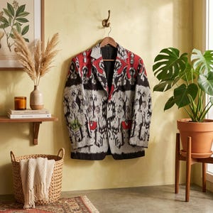 Könnte beinhalten: Ein langärmeliger Blazer mit einem auffälligen Ikat-Muster in Rot-, Schwarz- und Grautönen. Das Kleidungsstück verfügt über ein steigendes Revers, zwei Knöpfe auf der Vorderseite und zwei Pattentaschen. Der Stoff zeigt ein komplexes Muster.