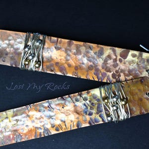 May include: A pair of rectangular copper earrings with a hammered, mottled finish in shades of brown, orange, and silver. Each earring features a section wrapped in thin silver wire with small metallic beads and a silver hook. Text reads Lost My Rocks.