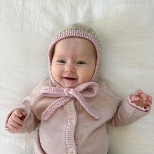 May include: A soft pink knitted cardigan with a button-down front and a matching bonnet featuring a pale green and pink striped pattern. The bonnet is secured under the chin with a knitted bow tie, and the cardigan has long sleeves with ruffled cuffs.