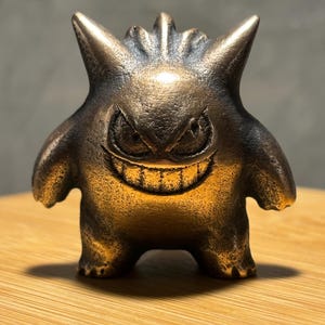 Könnte beinhalten: Eine kleine, metallische, bronzefarbene Figur eines runden, kreaturartigen Wesens mit spitzen Ohren, einem breiten, grinsenden Mund mit sichtbaren Zähnen und schmalen, bedrohlich wirkenden Augen. Die Oberfläche hat ein texturiertes Finish.