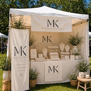 May include: A white outdoor vendor tent featuring the black logo MK MIKESGRAPHICSSHOP on the canopy, a vertical banner, and a table skirt. The display includes jewelry stands, white floral arrangements in pots, and small signs on a table.
