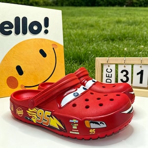 May include: A pair of red slip-on clogs featuring a Lightning McQueen design with the number 95 in yellow, white, and orange graphics on the side, blue and white eye details on the top, and a black and white mouth graphic on the front toe area.