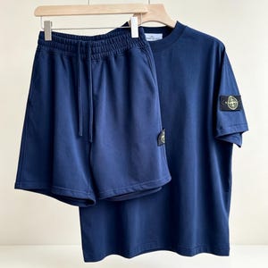 Peut inclure: Un t-shirt bleu marine à manches courtes et un short assorti à taille élastique avec cordon de serrage. Les deux pièces présentent un écusson rectangulaire noir sur la manche et la jambe, affichant un logo boussole jaune et vert.