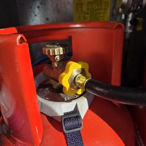 May include: A bright red propane gas cylinder featuring a brass valve assembly with a distinct yellow, flower-shaped plastic knob. A black rubber hose is securely attached to the valve, and a dark blue nylon strap is fastened around the white collar.