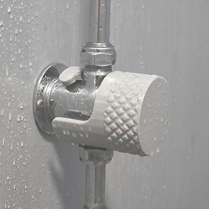 May include: A white cylindrical shower knob with a textured diamond pattern on the side, attached to a chrome metal pipe fixture. The knob is mounted on a circular chrome base against a light grey wall covered in water droplets.