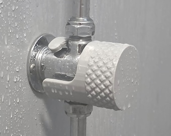 May include: A white cylindrical shower knob with a textured diamond pattern on the side, attached to a chrome metal pipe fixture. The knob is mounted on a circular chrome base against a light grey wall covered in water droplets.