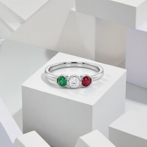 Op de afbeelding: Een zilverkleurige ring met drie ronde, in een gladde zetting geplaatste edelstenen die horizontaal zijn gerangschikt. De middelste steen is een heldere, gefacetteerde witte edelsteen, geflankeerd door een groene steen links en een rode steen rechts.