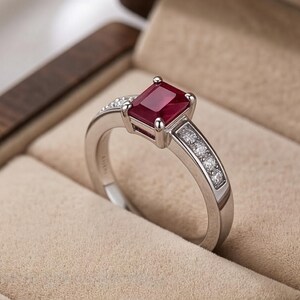 May include: A silver-toned ring featuring a rectangular, deep red gemstone set in the center. The band is accented with a channel setting of small, clear, round-cut stones along each side, leading up to the central ruby-colored stone.