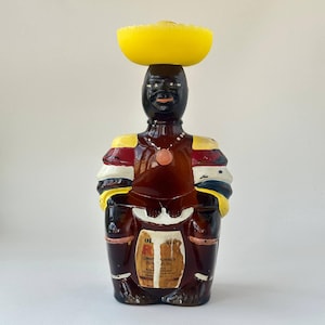 Op de afbeelding: Een bruine glazen karaf in de vorm van een persoon met een gele hoed, rood-gele mouwen en een ketting met een roze hanger. De figuur heeft een donkere huidskleur en een etiket op de voorzijde met de tekst Old Mr. Boston Limbo Rum 26 Fl. Oz.