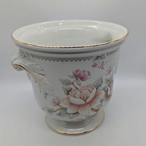 May include: A white ceramic planter featuring a delicate floral pattern with pink and peach blossoms, green leaves, and gold trim along the rim, base, and handles. The vessel has a flared top, a pedestal base, and two sculpted leaf-shaped side handles.
