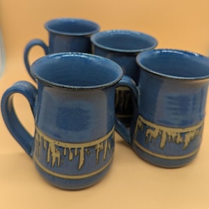 May include: A set of four blue ceramic mugs with a glossy finish and a dark rim. Each mug features a horizontal band near the base with a decorative drip pattern in a light tan color. The mugs have a curved handle and a wide, circular opening.