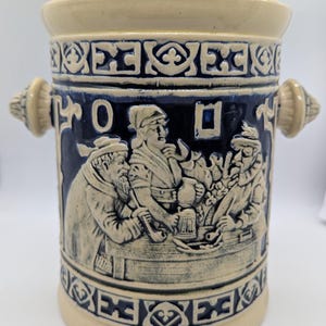 Könnte beinhalten: Ein zylindrischer Keramikbehälter mit Deckel in Creme und Kobaltblau. Die Außenseite zeigt ein Relief mit drei Personen an einem Tisch, umrahmt von dekorativen geometrischen Mustern und zwei seitlichen Griffen.