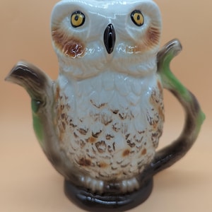 Puede incluir: Una tetera de cerámica con forma de búho, con plumas blancas, manchas marrones y ojos amarillos. El asa y el pico están diseñados para parecer ramas de árbol con hojas verdes, y el búho está posado sobre una base de color marrón oscuro.