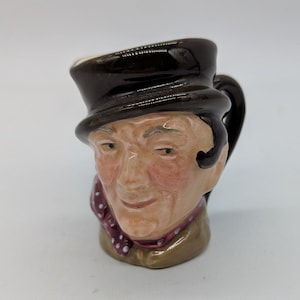 May include: A ceramic character jug shaped like a person's head wearing a dark brown hat. The figure has a light skin tone, a maroon scarf with white polka dots around the neck, and a tan collar. The handle is attached to the back of the head.