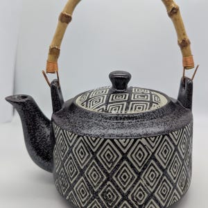 Può includere: Una teiera in ceramica con finitura scura maculata e un motivo geometrico a rombi bianchi. La teiera presenta un beccuccio curvo, un coperchio abbinato con pomello e un manico curvo in bambù fissato con fili di rame su entrambi i lati.