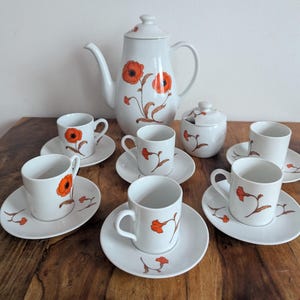 Op de afbeelding: Een wit porseleinen theeservies bestaande uit een hoge theepot, een suikerpot met deksel en vijf kopjes met bijpassende schoteltjes. Elk onderdeel is versierd met een handgeschilderd oranje klaproosmotief met bruine stengels en bladeren.