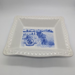 May include: A square white ceramic dish with a raised, textured border pattern. The center features a blue illustration of a 19th-century wheat harvest scene with a horse-drawn cart and the text 1892 1992 SHREDDED WHEAT in a circular emblem.