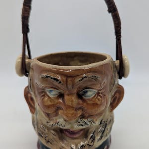 May include: A ceramic character mug shaped like a bearded man's head with a glossy finish, featuring painted facial details, blue eyes, and a dark brown woven handle attached to the sides with two cream-coloured circular accents at the top rim.