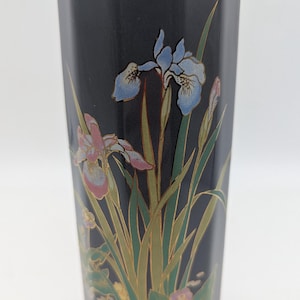 Op de afbeelding: Een hoge, zeshoekige zwarte vaas met een geschilderd bloemmotief van blauwe en roze irissen met groene stengels en bladeren. De rand van de vaas is afgewerkt met een dunne goudkleurige metalen band, wat een contrast vormt met het donkere oppervlak.