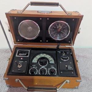 Könnte beinhalten: Ein hölzerner Radiowecker mit Metallgriff und Eckbeschlägen. Der obere Teil enthält einen runden Lautsprecher und ein analoges Ziffernblatt. Das Bedienfeld umfasst eine Frequenzanzeige, einen Netzschalter, Lautstärke- und Klangregler sowie eine Schlummertaste.