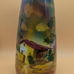 May include: A tapered glass vase featuring a hand-painted landscape scene with a small house, trees, and a golden-brown ground. The upper portion of the vase transitions into a deep blue sky, and the rim is finished with a thin gold-colored band.