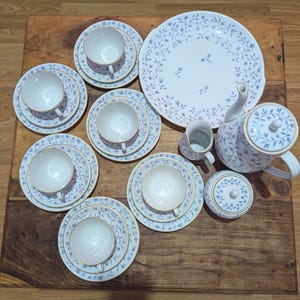 Puede incluir: Un juego de té de porcelana blanca con un delicado patrón de enredaderas y hojas azules con borde dorado. El conjunto incluye una bandeja grande, una tetera con tapa, una jarra de leche, un azucarero con tapa y cinco tazas con sus platos.