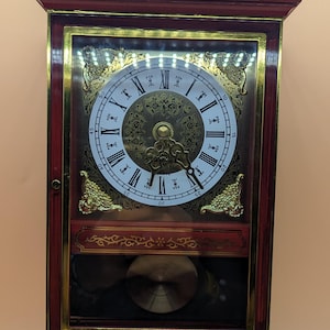 May include: A wall clock with a dark red wooden frame and gold accents. The clock face features a white circular dial with black Roman numerals, ornate gold hands, and a gold pendulum visible behind a glass panel. The top and bottom are decorated with gold filigree patterns.