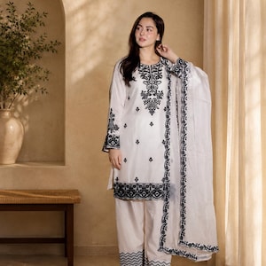 Op de afbeelding: Een wit driedelig ensemble bestaande uit een tuniek met lange mouwen, een wijde broek en een bijpassende sjaal. De tuniek is voorzien van gedetailleerd zwart borduurwerk bij de halslijn, manchetten en zoom, met zwarte randen op de broek en sjaal.