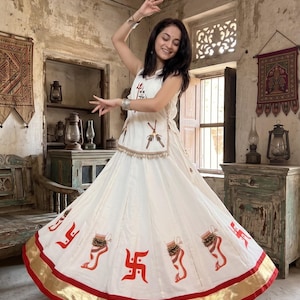 Op de afbeelding: Een wit mouwloos topje en een lange, uitlopende rok met een goudkleurige en rode zoom. De rok is versierd met oranje geschilderde motieven van voeten met enkelbandjes en rode swastika-symbolen. De top heeft een rode drietand-afbeelding.