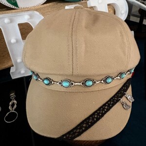 May include: A tan newsboy cap featuring a silver-toned chain embellished with oval turquoise-coloured stones encircling the crown. A black lace ribbon is wrapped diagonally across the brim, accented with two small silver-toned leaf-shaped charms.