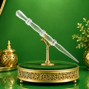 Puede incluir: Un objeto transparente en forma de varita con una punta afilada y un mango texturizado, montado sobre un soporte de metal dorado. El soporte cuenta con un brazo central y una base circular con detalles florales y una superficie de terciopelo.