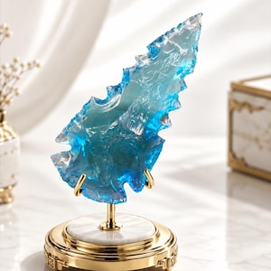 Könnte beinhalten: Eine durchscheinende, pfeilspitzenförmige Skulptur in Blau mit gezackten, texturierten Kanten. Das Objekt ist auf einem goldfarbenen Metallständer mit kreisförmigem Sockel und weißer Marmoroptik montiert und wird von zwei kleinen goldenen Halteklammern gestützt.