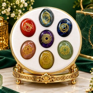 Könnte beinhalten: Ein runder weißer Präsentationsständer mit goldenem Metallrahmen enthält sieben ovale, polierte Edelsteine. Jeder Stein ist mit einem goldenen Chakra-Symbol graviert. Die Steine sind kreisförmig angeordnet, mit einem violetten Stein in der Mitte und sechs weiteren Steinen in Rot, Braun, Gelb, Grün, Blau und Dunkelgrau.