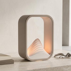 May include: A rectangular table lamp with rounded corners and a matte taupe exterior frame. The interior features a layered, white, wave-like structure that glows with a warm light, creating a mountain-inspired silhouette within the open center.