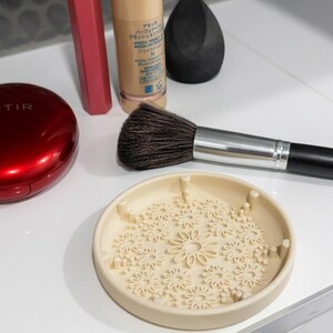 Op de afbeelding: Een beige ronde siliconen make-up kwastenreiniger mat met een verhoogd bloemenpatroon, geplaatst naast een zwarte make-up spons, een make-up kwast met een zwarte steel, een beige fles, een rode lippenstift en een rood compact doosje.