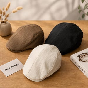 May include: Three flat caps in solid colors: one brown, one black, and one off-white. Each cap features a rounded crown, a short stiff brim, and a textured fabric construction. The caps are arranged together on a flat surface.