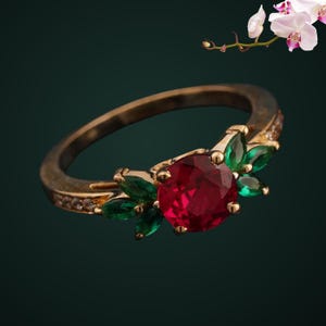 May include: A gold ring featuring a central round-cut red gemstone flanked by marquise-cut green stones arranged in a leaf-like pattern. The band is crafted from gold metal and includes small clear stones embedded along the sides of the ring shank.