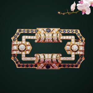 Può includere: Una spilla rettangolare in oro con un design geometrico traforato. Il pezzo è incastonato con numerose pietre bianche trasparenti e accentuato da pietre blu scuro lungo il bordo esterno, creando un aspetto strutturato e simmetrico.