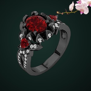 May include: A black metal ring featuring a large oval-cut deep red center stone, accented by smaller heart-shaped red stones and numerous tiny clear white stones embedded along the band and the setting surrounding the central gemstone.