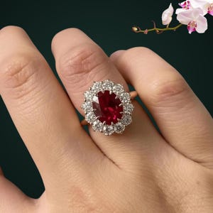 May include: A ring featuring a large, oval-cut deep red ruby at the center, surrounded by a halo of twelve round, brilliant-cut white diamonds set in a metallic band. The design showcases a classic cluster arrangement with a polished finish.