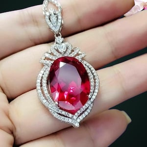 May include: A silver-toned pendant featuring a large, oval-cut, deep red gemstone as the centerpiece. The stone is surrounded by a decorative frame encrusted with numerous small, clear, sparkling stones. A matching stone-set bail sits atop.