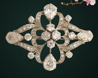 May include: A silver-toned brooch featuring an intricate openwork design set with numerous clear, faceted stones. The piece includes four prominent pear-shaped stones at the cardinal points and a central cluster of round stones within a frame.