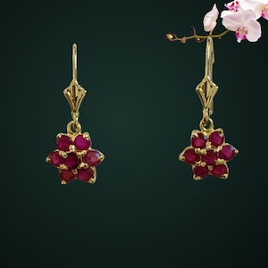 May include: A pair of gold drop earrings featuring a flower-shaped cluster of six round, deep pinkish-red gemstones. Each earring is suspended from a gold lever-back closure, showcasing a symmetrical floral design with polished metal settings.